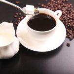 Een kop koffie waar een suikerklontje in wordt gedaan, naast het kopje staat een kannetje melk en op de achtergrond liggen koffiebonen